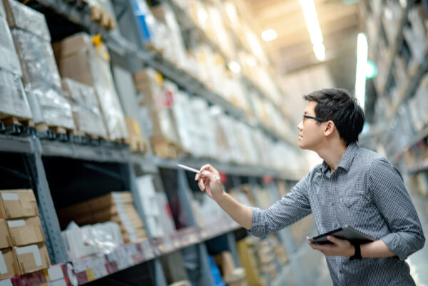 Young Asian man worker doing stocktaking of product in cardboard box on shelves in warehouse by using digital tablet and pen. Physical inventory count concept