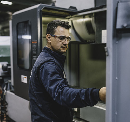 Portrait of apprentice engineering worker young man working, examining and operating CNC plastic injection molding machinery in factory warehouse after studied manufacturing apprenticeship program certifies